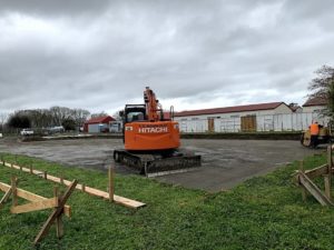 A construction site with a large orange Hitachi excavator parked on a gravel area. Wooden frames are in the foreground, with grassy areas and buildings in the background under a cloudy sky.