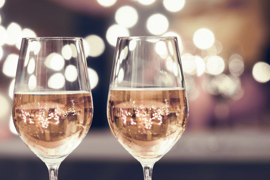 Two wine glasses filled with champagne are in focus, with a backdrop of blurred, warm white bokeh lights creating a festive atmosphere.