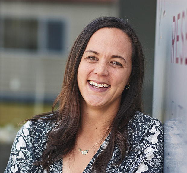 A person with long dark hair is smiling broadly while leaning against a wall. They are wearing a patterned top and have a necklace. The background is slightly blurred, showing a building and window.