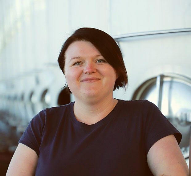 A woman with short dark hair and a navy shirt smiles at the camera, standing in front of large, shiny metal tanks, possibly at a brewery or industrial facility.