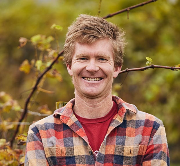 Matthew Corbett A smiling man with short, tousled blonde hair wearing a red t-shirt and a plaid flannel shirt stands outdoors in front of a blurred background of autumn leaves and branches.