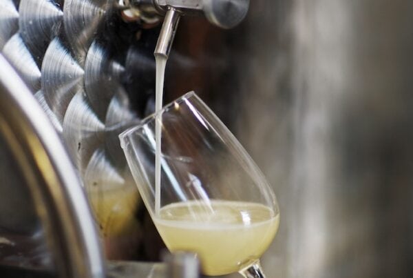A person holds a glass at an angle, collecting cloudy white wine from a stainless steel spout attached to a large metal fermenting tank.