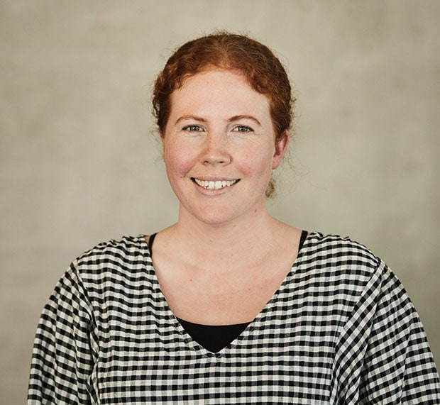 A woman with curly red hair tied back, wearing a black and white checkered shirt, smiles at the camera in front of a plain beige background.