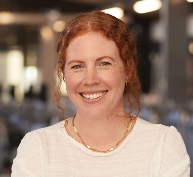 A woman with curly red hair tied back, wearing a black and white checkered shirt, smiles at the camera in front of a plain beige background.