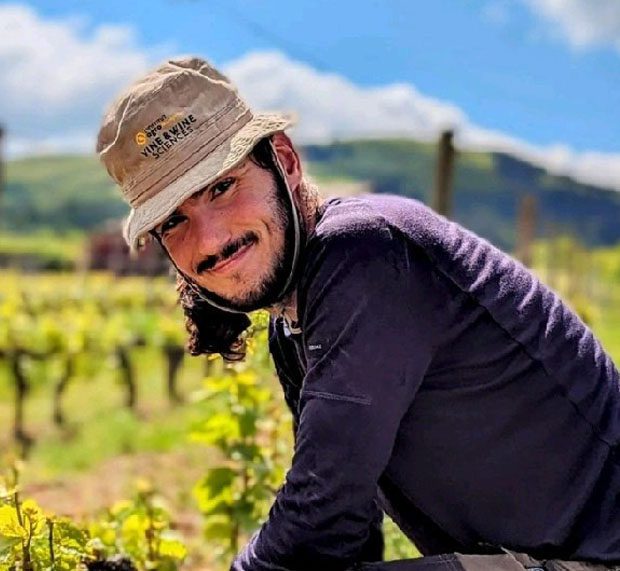 A man in a wide-brim hat and dark shirt smiles while crouching among green plants in a vineyard, with blurred hills and blue sky in the background.