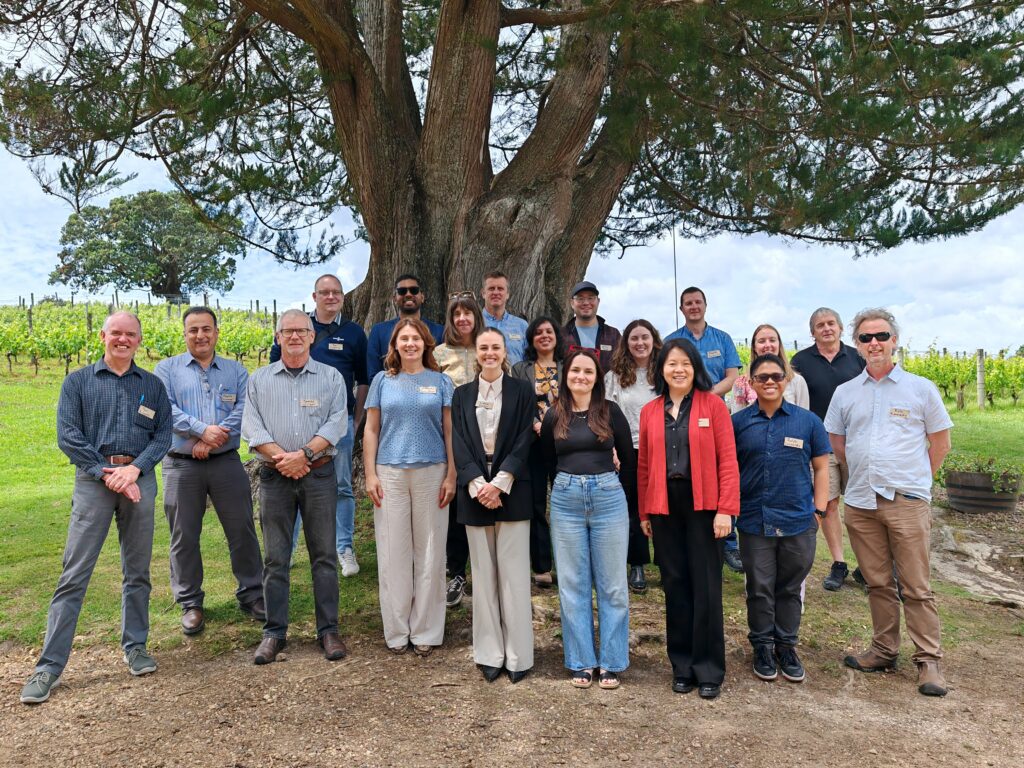 A group of 17 people pose for a photo outdoors in front of a large tree, with green grass and vineyard rows in the background. Most are smiling and wearing name badges.