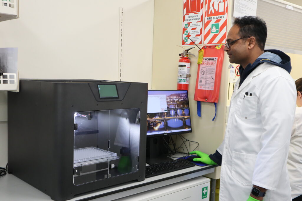 A person in a lab coat and green gloves stands next to a 3D printer and a computer monitor in a laboratory, with safety signs and a fire extinguisher visible on the wall behind them.