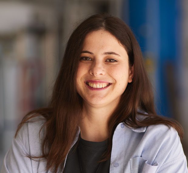 A young woman, long brown hair, in a light blue shirt over a black top, smiles at the camera; background blurred in blue tones.