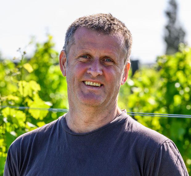 [Name] wears a dark T-shirt and smiles outdoors among lush green vineyard plants on a sunny day.