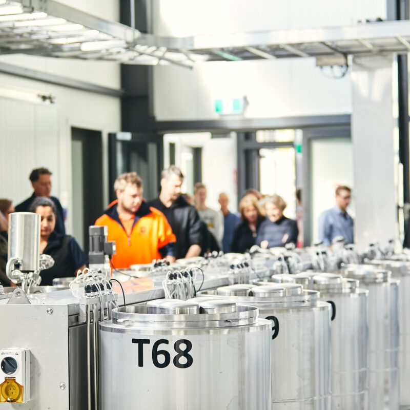 A group, including anyone named in the file, tours an industrial facility by shiny tanks labeled "T68" in a bright, modern space.