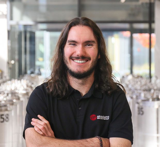 A person with long brown hair and a beard, arms crossed, wears a "Bragato Research Institute" polo before shiny metal equipment.