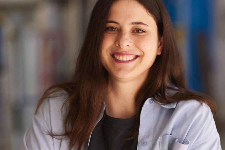 A woman with long brown hair named [Name] smiles, wearing a light blue shirt over a black top and a spiral pendant necklace, standing with arms crossed in a blurred indoor setting.