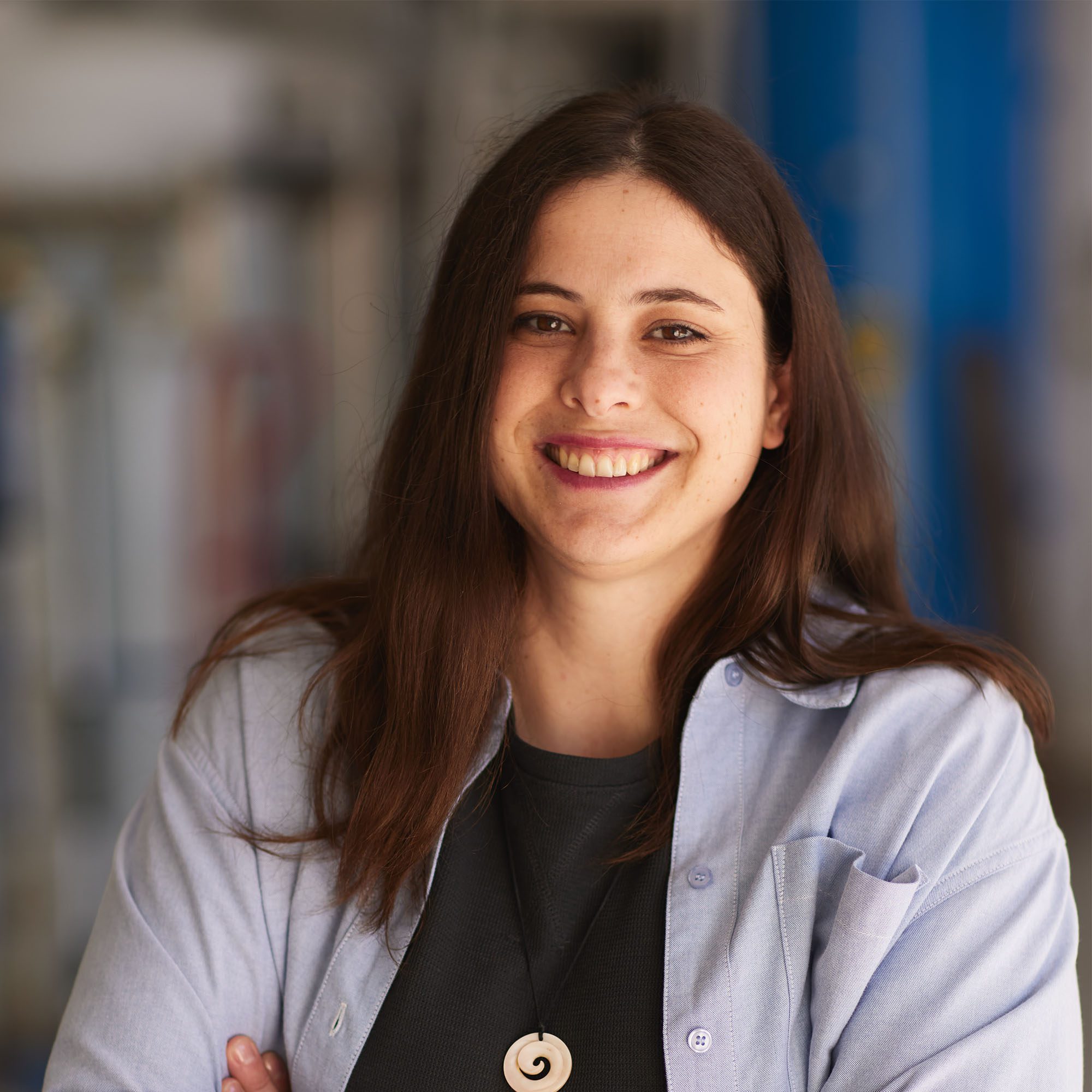 A woman with long brown hair named [Name] smiles, wearing a light blue shirt over a black top and a spiral pendant necklace, standing with arms crossed in a blurred indoor setting.