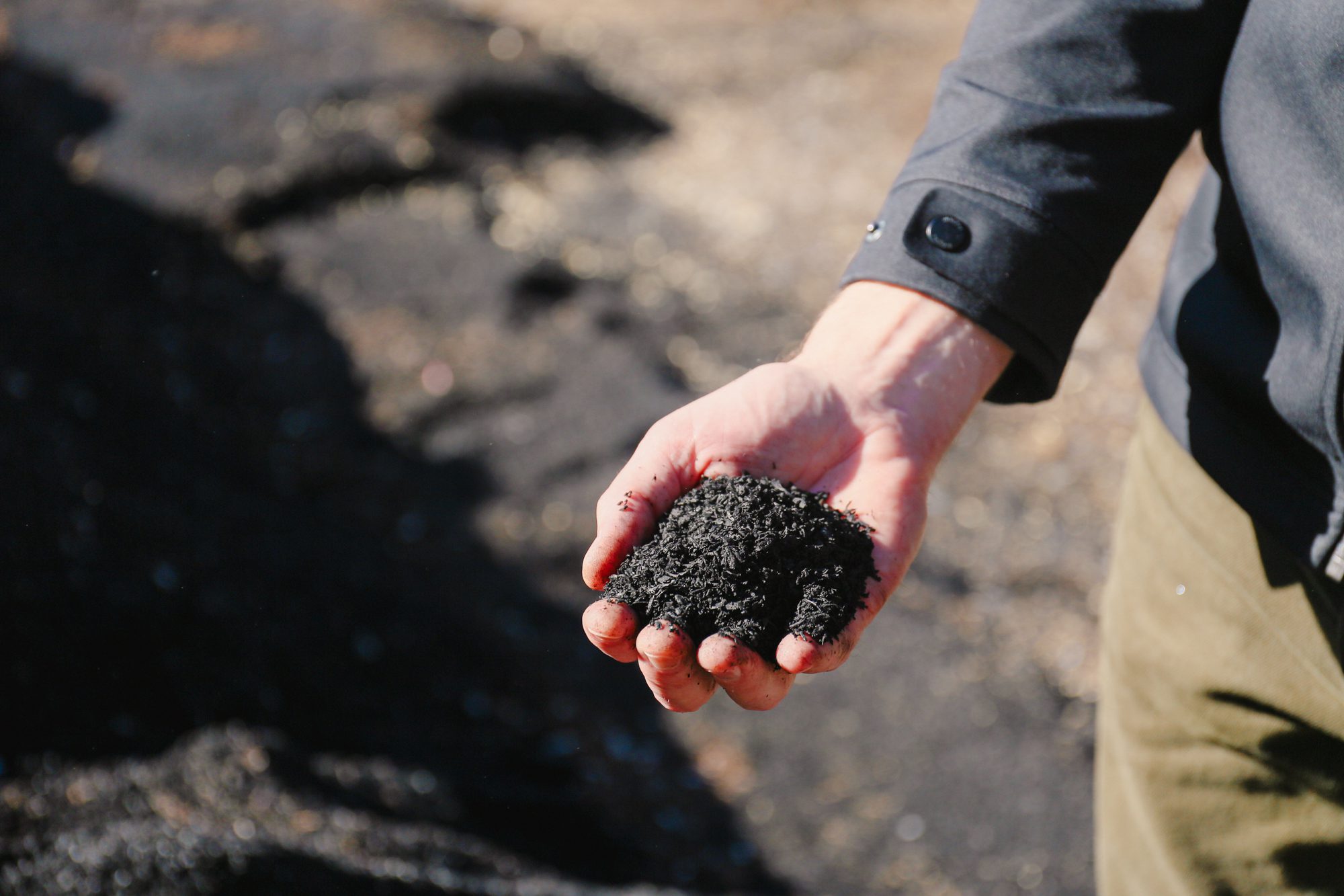 A person in a dark sleeve and green pants holds a handful of black soil or compost, with additional soil visible in the softly blurred background.
