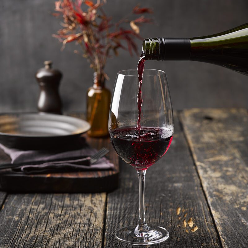 Red wine being poured into a glass on a rustic wooden table, surrounded by a plate, napkin, cutlery, pepper grinder, and a dried plant in the background.