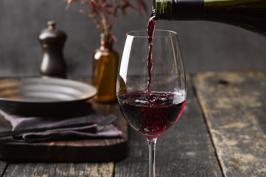 Red wine being poured into a glass on a rustic wooden table, surrounded by a plate, napkin, cutlery, pepper grinder, and a dried plant in the background.